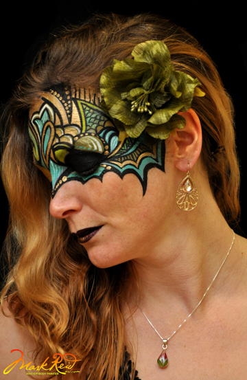 woman with the top of her face pained in a mask that's blue and gold with a green flower behind her ear